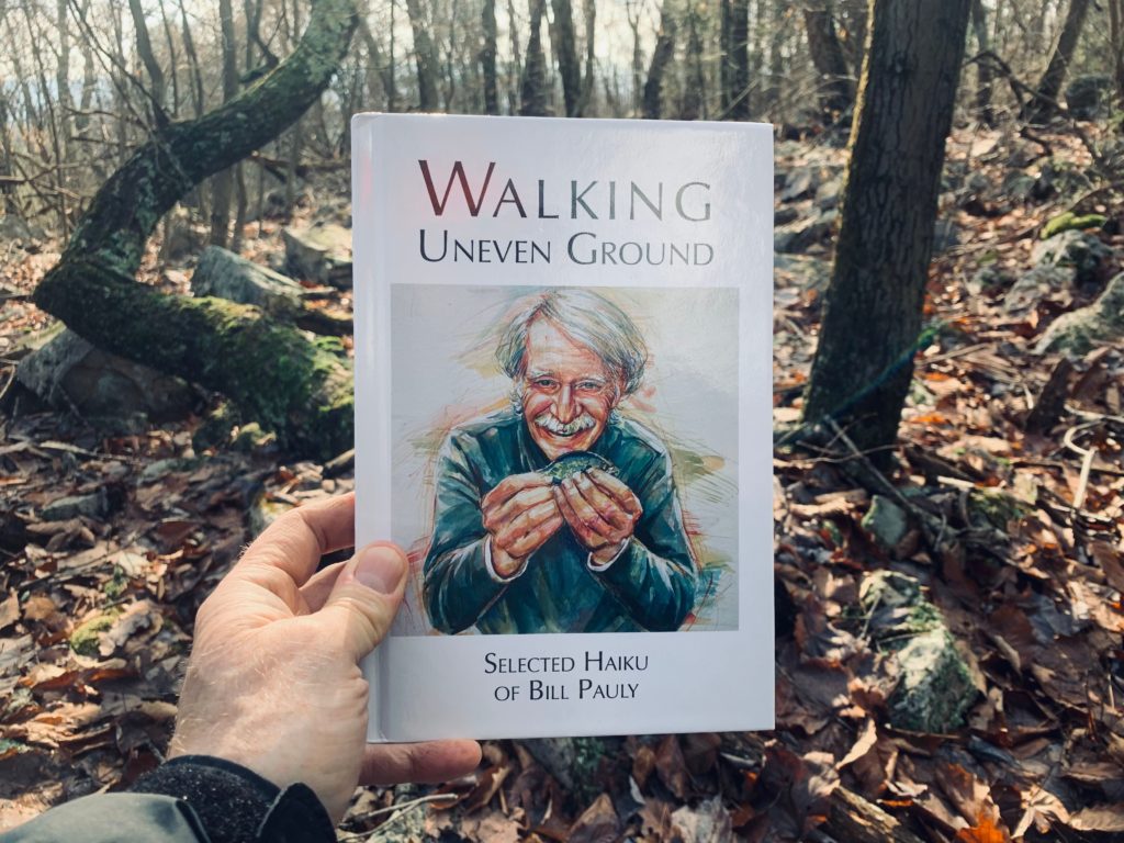 A hand holds up the cover of "Walking Unever Ground:  Selected Haiku of Bill Pauly" in a late-autumn forest